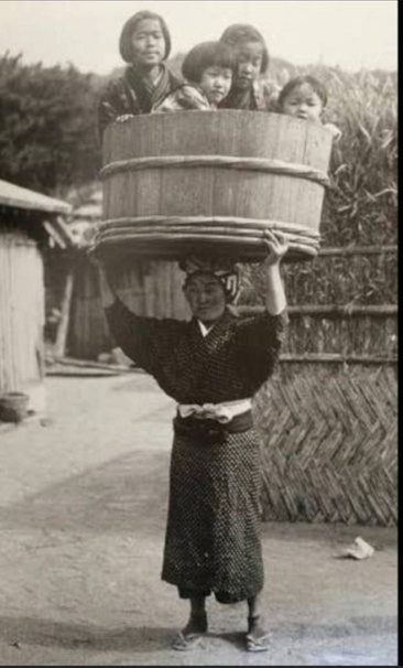 An archival photograph from Japan: a woman carrying a large wooden tub on her head with three children sitting inside it.