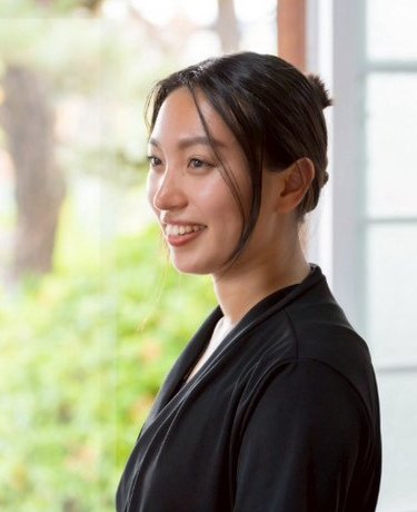 Portrait of Akari Mukai, smiling, in a softly lit room with greenery behind her.